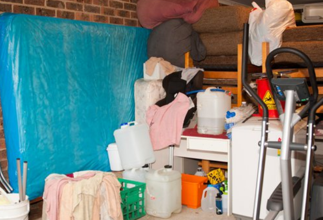 A cluttered garage packed with a blue-wrapped mattress, plastic containers, towels and an exercise machine during a hoarder clearance.