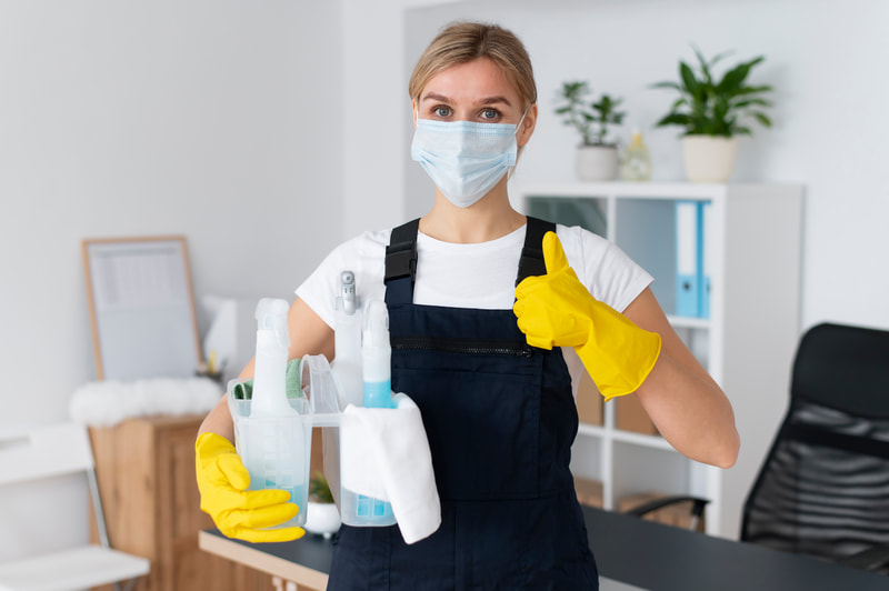 A cleaner wearing a face mask and yellow gloves holds cleaning spray bottles and gives a thumbs up in an office.