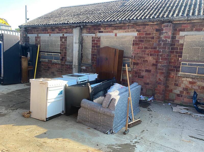 A pile of old furniture, a cooker and a wardrobe stacked outside a brick building ready for clearance.