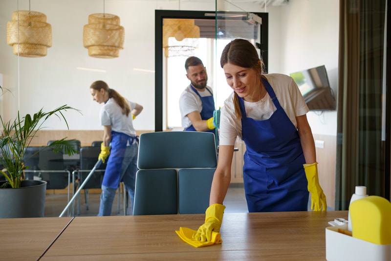Three cleaners in blue aprons wipe down a wooden table and mop the floor of an office space. Three cleaners in blue aprons wipe down a wooden table and mop the floor of an office space.