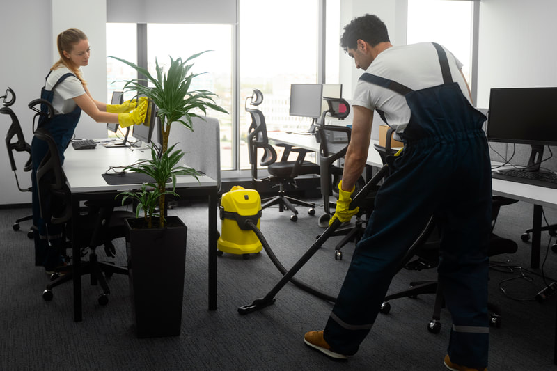 Two cleaners in navy overalls wipe down office desks and vacuum the carpet between workstations. Two cleaners in navy overalls wipe down office desks and vacuum the carpet between workstations.